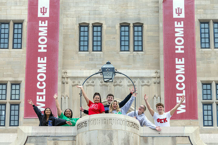 Students wearing Greek letter sweatshirts stand with their arms in the air on a balcony with banners that say, "Welcome Home."